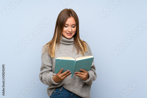 Teenager girl with sweater over isolated blue background holding and reading a book