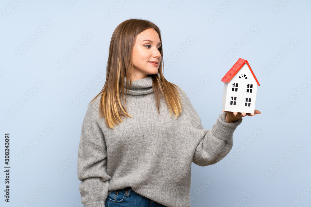 Teenager girl with sweater over isolated blue background holding a little house Teenager girl with sweater over isolated blue background holding a little house