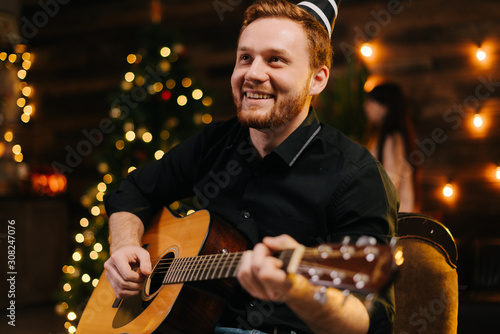 Laughing young man is playing guitar. Guy is looking happily and carefree. Man in festive hat celebrating Christmas or new year.