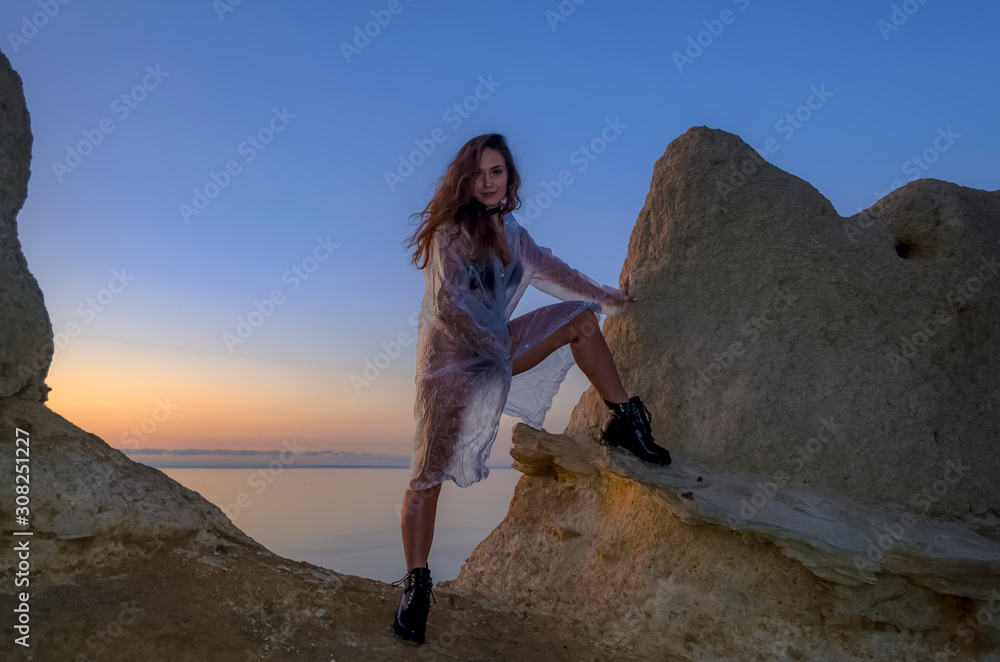 Naklejka premium Silhouette of a young and sexy girl. She stands on a mountain and poses, dressed in a cloak and black boots against the sunset. Happy celebration victory success woman