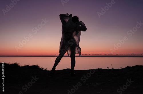 Silhouette of a young and sexy girl. She stands on a mountain and poses, dressed in a cloak and black boots against the sunset. Happy celebration victory success woman
