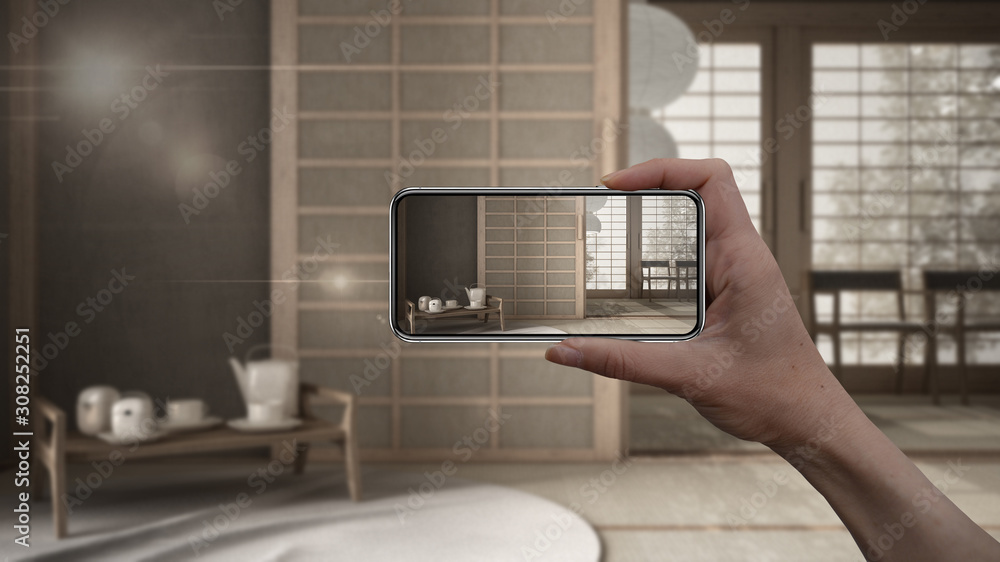 Hand holding smart phone, AR application, simulate furniture and interior design products in real home, architect designer concept, blur background, empty room with futon and tatami Hand holding smart phone, AR application, simulate furniture and interior design products in real home, architect designer concept, blur background, empty room with futon and tatami