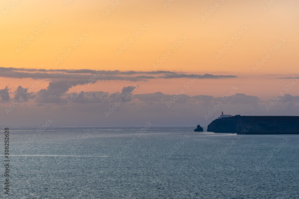 The landscape is warm colours. A Lighthouse on a rock against the backdrop of a sunset sky.