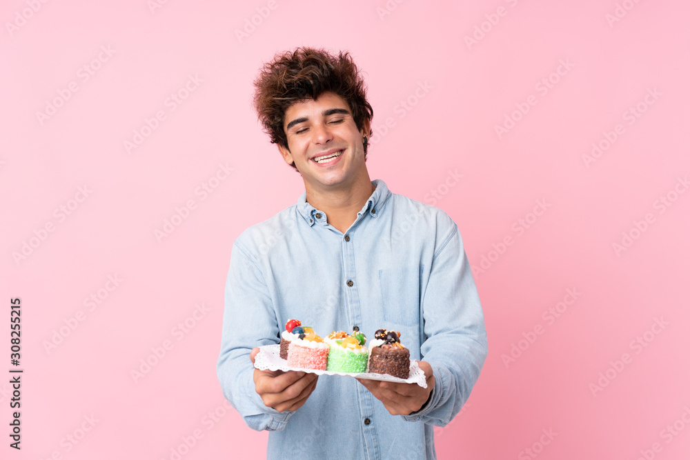 Young caucasian man over isolated pink background holding mini cakes Young caucasian man over isolated pink background holding mini cakes
