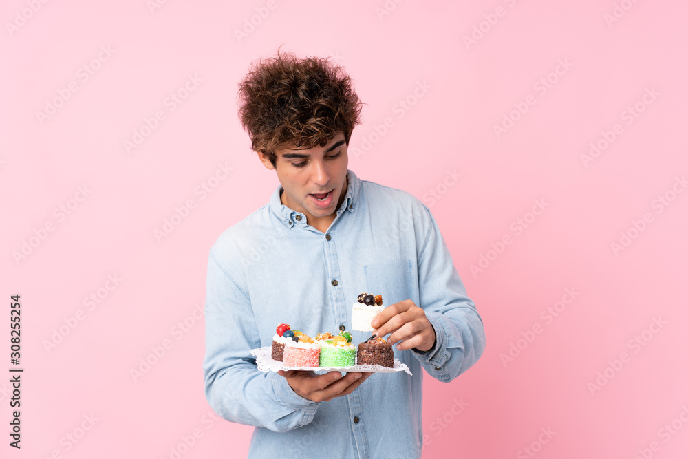 Young caucasian man over isolated pink background holding mini cakes and surprised Young caucasian man over isolated pink background holding mini cakes and surprised