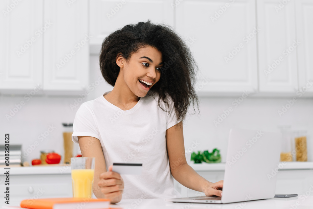 Excited African American woman ordering food online. Beautiful hipster girl with emotional face using credit card and laptop for online shopping, making transaction, standing on white kitchen at home Excited African American woman ordering food online. Beautiful hipster girl with emotional face using credit card and laptop for online shopping, making transaction, standing on white kitchen at home