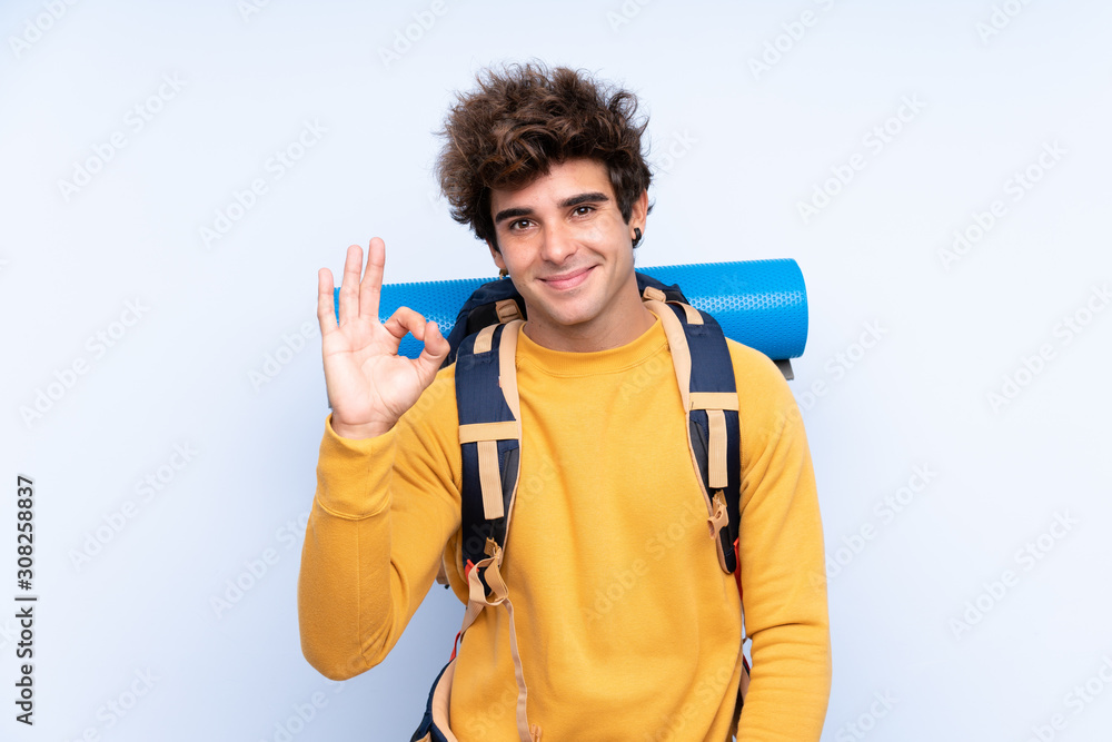 Young mountaineer man with a big backpack over isolated blue background showing an ok sign with fingers Young mountaineer man with a big backpack over isolated blue background showing an ok sign with fingers