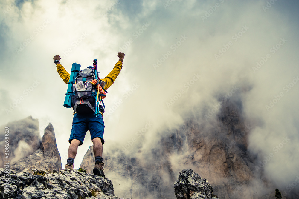 Obraz premium Young man hiker standing on stone mountain with raised up arms with cloudy sky and fog. Yellow jacket, backpack, black beard and beanie. Traveling Dolomites, Italy.