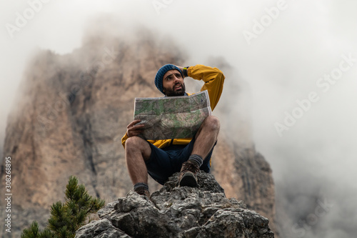 Young man hiker sitting on stone mountain reading map, with cloudy sky and fog. Yellow jacket, backpack, black beard and beanie. Traveling dolomites, Italy. photo