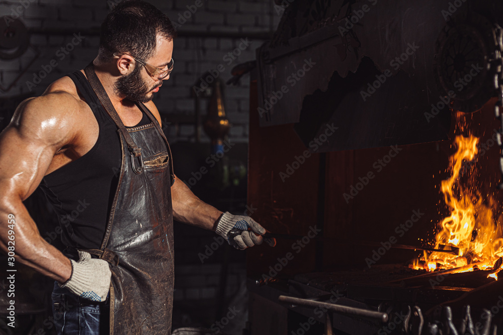 Young caucasian man blacksmith, strong iron worker in workshop, work place, isolated dark space with furnace