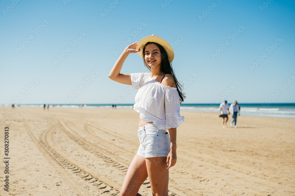 joyful girl on the beach is standing in shorts and a blouse holding her hat on her hand and smiling cute against the background of the oceans