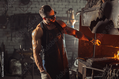 muscular adult with beard wearing leather apron working in workshop, heating. hardened metal in furnace