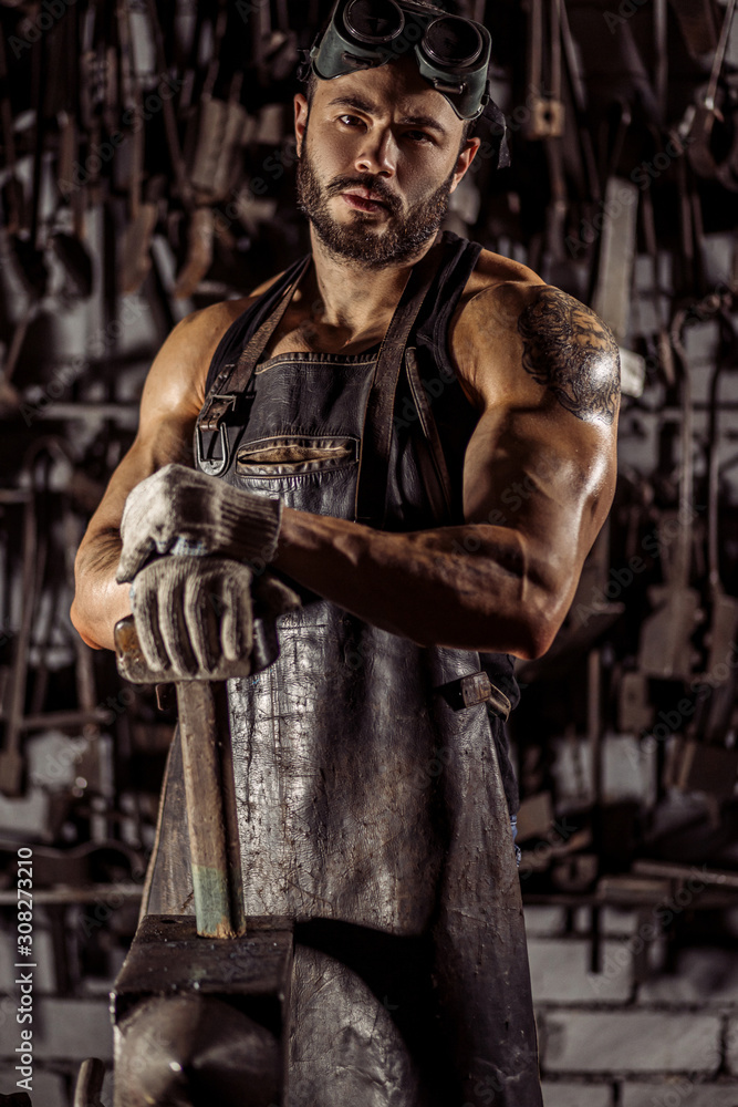 portrait of young muscular strong caucasian man blacksmith looking at camera, wearing leather apron uniform, holding hammer isolated in dark space, workshop
