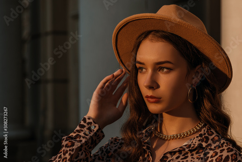 Outdoor close up fashion portrait of young elegant model, woman wearing beige hat, golden earrings, trendy chain necklace, posing in street of European city, at sunset. Copy, empty space for text