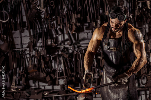 strong muscular brutal confident blacksmith man shaping red hot metal with hammer isolated in workshop, wearing leather apron, dark space