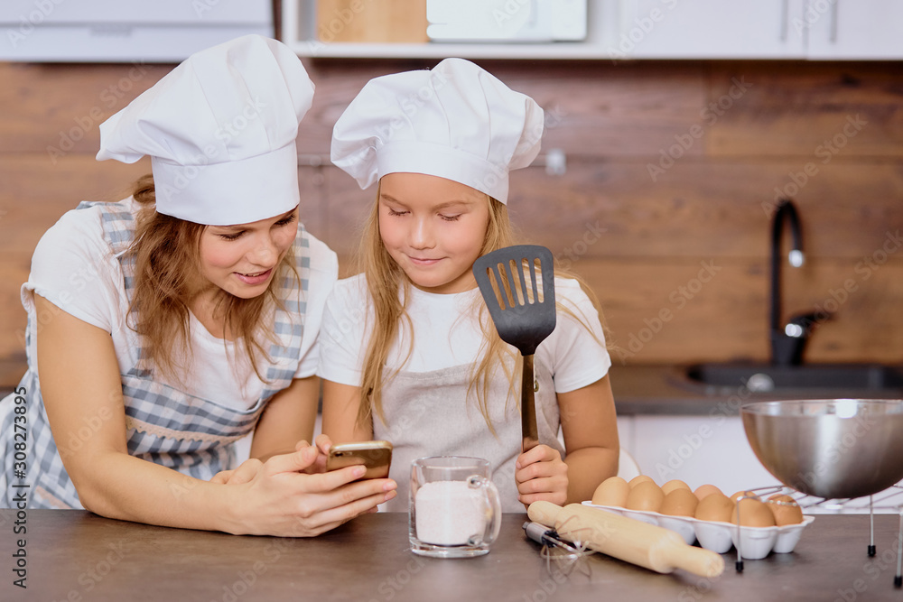 caucasian mother and kid daughter in caps and aprons look at screen of smartphone watching delicious cake recipe, going to cook together caucasian mother and kid daughter in caps and aprons look at screen of smartphone watching delicious cake recipe, going to cook together