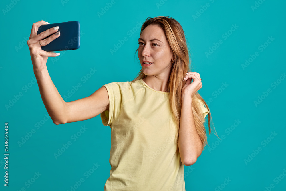 Beautiful girl portrait with half length phones on a blue background. A young woman takes a selfie and speaks on the phone. Beautiful girl portrait with half length phones on a blue background. A young woman takes a selfie and speaks on the phone.