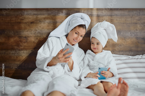 lovely female and her daughter sit on bed in bathrobes and towel gusing smartphone, mobile phone, digital device