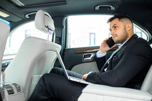 Handsome bearded man working on laptop while talking on phone in his luxurious car, side view © alfa27