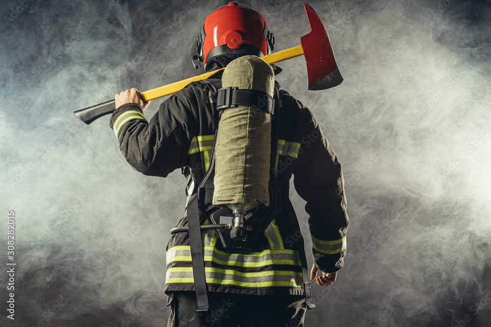 Fototapeta premium rear view on fire fighter stand holding hammer isolated over smoky background, wearing red helmet