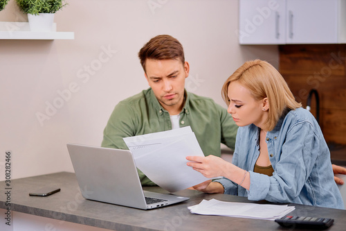 young caucasian woman and man wearing casual wear checking financial situation in family, doing invoice verficitaion using laptop, papers and calculator