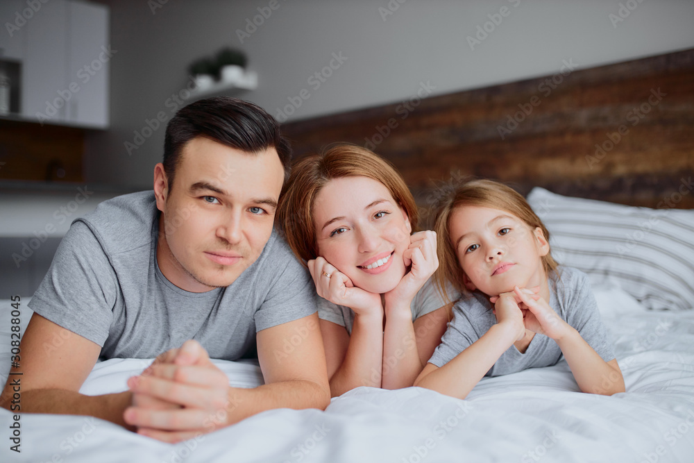 portrait of beautiful happy family lying on bed together, mother father and kid girl kissing hugging and laughing. indoors