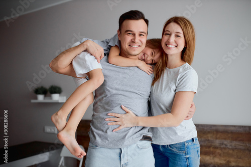 portrait of beautiful young caucasian family, parents with daughter, posing at camera isolated in bedroom, hug each other