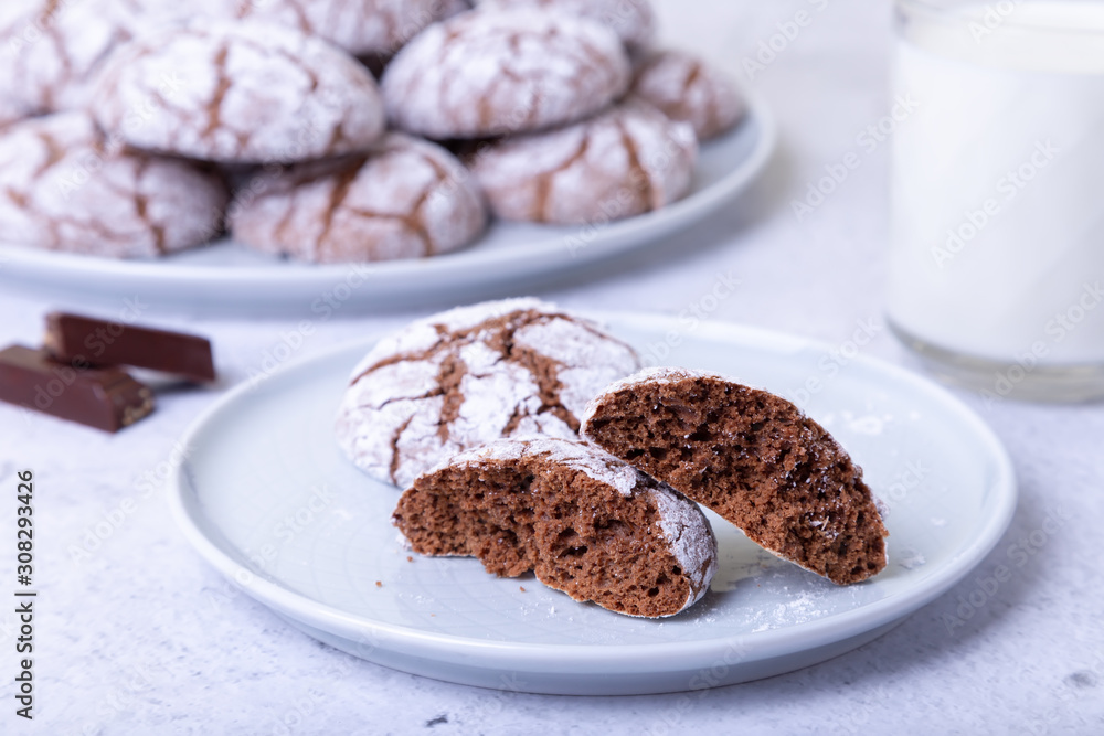 Chocolate cookies with cracks, strewed with icing sugar. Homemade baking. Background: a cup of milk, chocolate and a plate of cookies. Selective focus, close-up. Chocolate cookies with cracks, strewed with icing sugar. Homemade baking. Background: a cup of milk, chocolate and a plate of cookies. Selective focus, close-up.