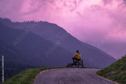 Young man sitting on a bench in front of the mountains after sunset with purple sky. Dolomites,South Tyrol, Italy © WineDonuts
