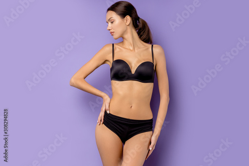 portrait of beautiful slim fit and sexy woman wearing black underwear standing isolated over purple background, looking side.
