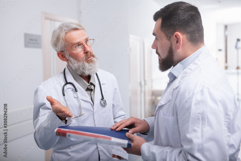 Senior doctor talking to his younger intern. Two male doctors chatting in the hall of a hospital Senior doctor talking to his younger intern. Two male doctors chatting in the hall of a hospital
