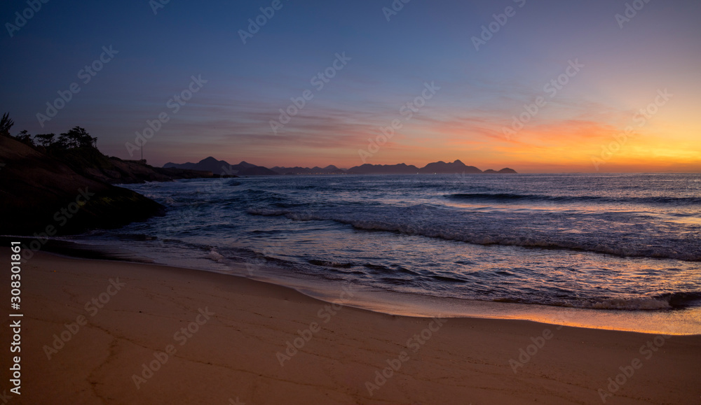 Warm colourful glowing sunrise at the crack of dawn at the Arpoador Devil's beach in Rio de Janeiro with waves coming in reflecting the orange colours of the sun and a pristine sand beach
