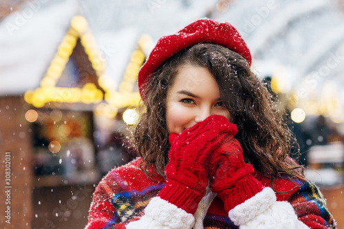 Festive Christmas fair, winter holidays concept: happy smiling woman wearing red beret, scarf, mittens warming her hand, posing at festive street market. Copy, empty space for text