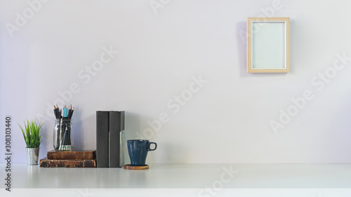 Comfortable workspace with books, pencil, coffee, plant and photo frame on white table and white wall.