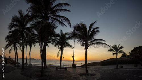 Panorama of a sunrise on the Arpoador boulevard with the Devils beach and silhouetted palm trees in Rio de Janeiro against a clear orange and blue sky © Maarten Zeehandelaar