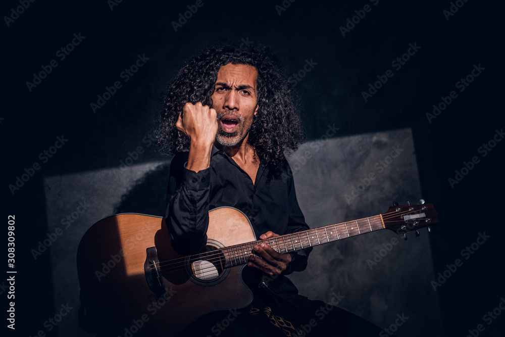 Obraz premium Charismatic rock singer is posing with his guitar at dark photo studio and showing a fist.