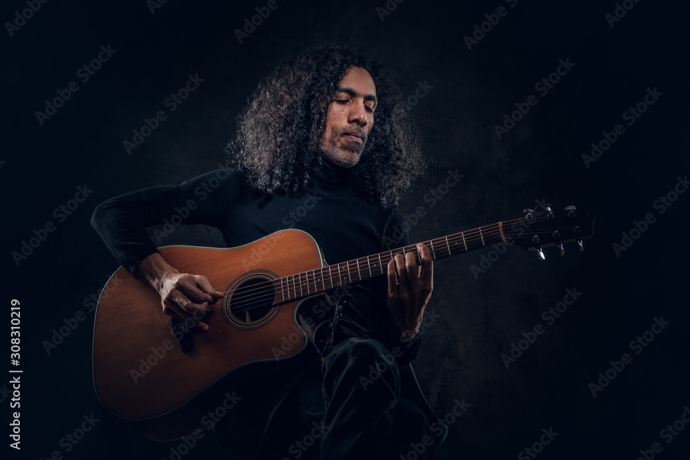Obraz premium Handsome man with long hair is playing acoustic guitar at dark studio.