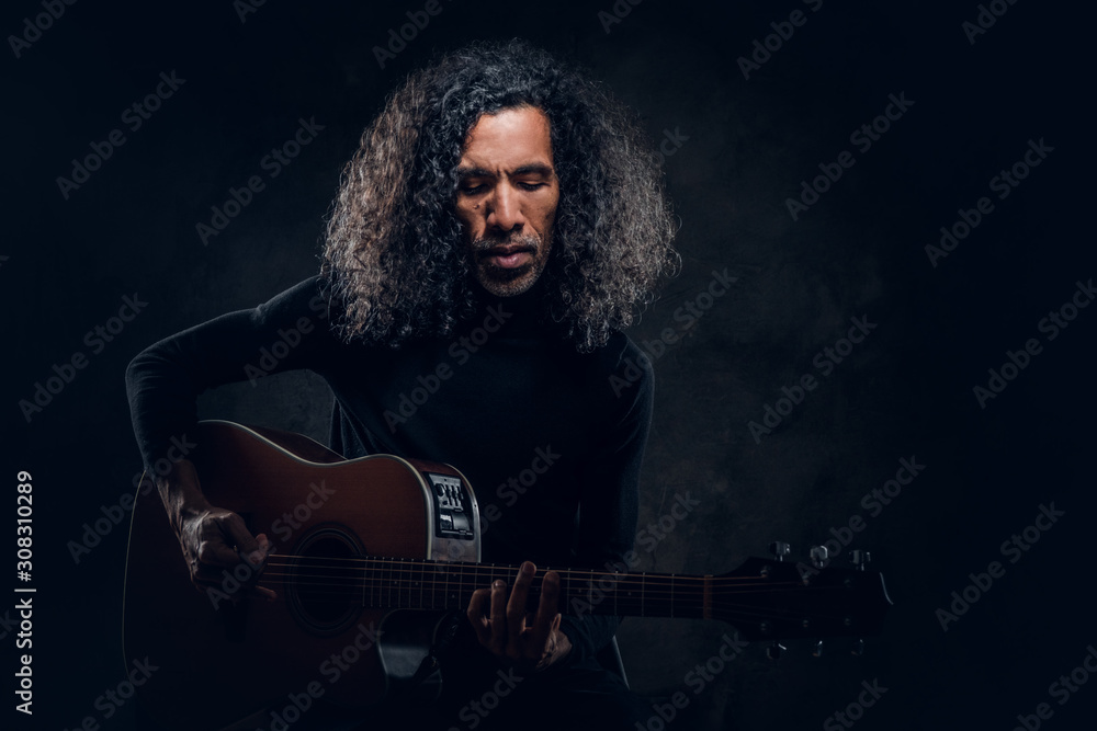 Fototapeta premium Handsome man with long hair is playing acoustic guitar at dark studio.