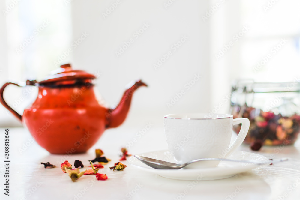 View of cup of tea, red teapot, herbal and infusion elements in a white room. Breakfast, food, and wellness concept. View of cup of tea, red teapot, herbal and infusion elements in a white room. Breakfast, food, and wellness concept.
