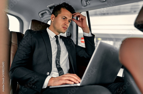 Young handsome businessman is sitting in luxury car. Serious man in suit is working with laptop while being in trip. © Valerii Apetroaiei