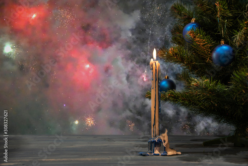 Christmas tree with decorations and a candle on a wooden table against the background of the night sky with firework.