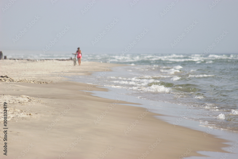 A girl goes next to a bicycle along the beach against the background of the sea. In the distance, a woman walks in the fresh air of the sea beach.