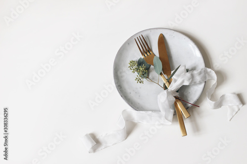 Festive table setting. Golden cutlery, berry eucalyptus branch, porcelain plate and silk ribbon isolated on white table background. Mediterranean wedding or restaurant menu concept. Flat lay, top view