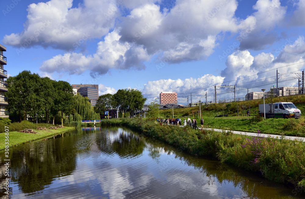 Amsterdam, Holland, August 2019. On the outskirts the canals are lined only with trees and greenery. The foliage is reflected in the water. More distant from the shore modern buildings. People walk