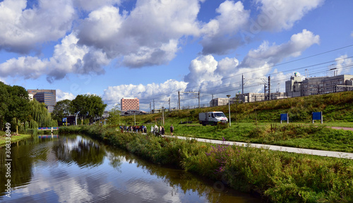 Amsterdam, Holland, August 2019. On the outskirts the canals are lined only with trees and greenery. The foliage is reflected in the water. More distant from the shore modern buildings. People walk