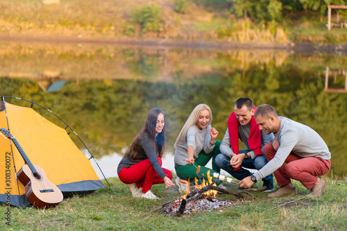 travel, tourism, hike, picnic and people concept - group of happy friends frying sausages on campfire near lake. © Kate