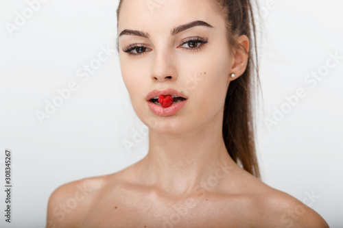 Portrait of a young girl with ponytail  with naked shoulders  holds in the mouth a raspberry  isolated on a white background. Concept about youth  young skin  eyebrows  eyelashes  beauty