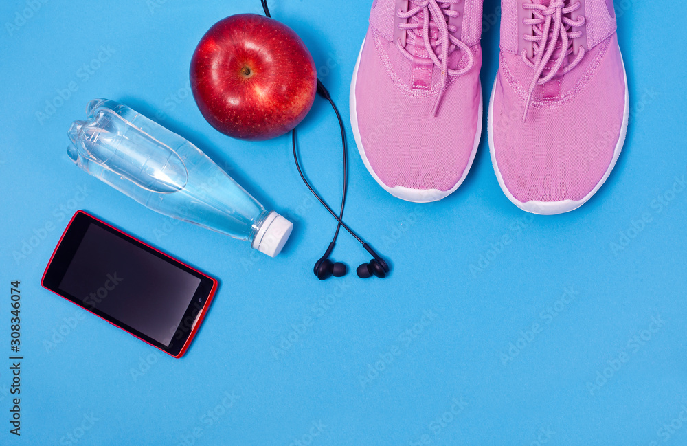 Pink sport shoes, earphones, red apple, smartphone and bottle of water on a blie background. Concept healthy lifestyle, sport and diet. 