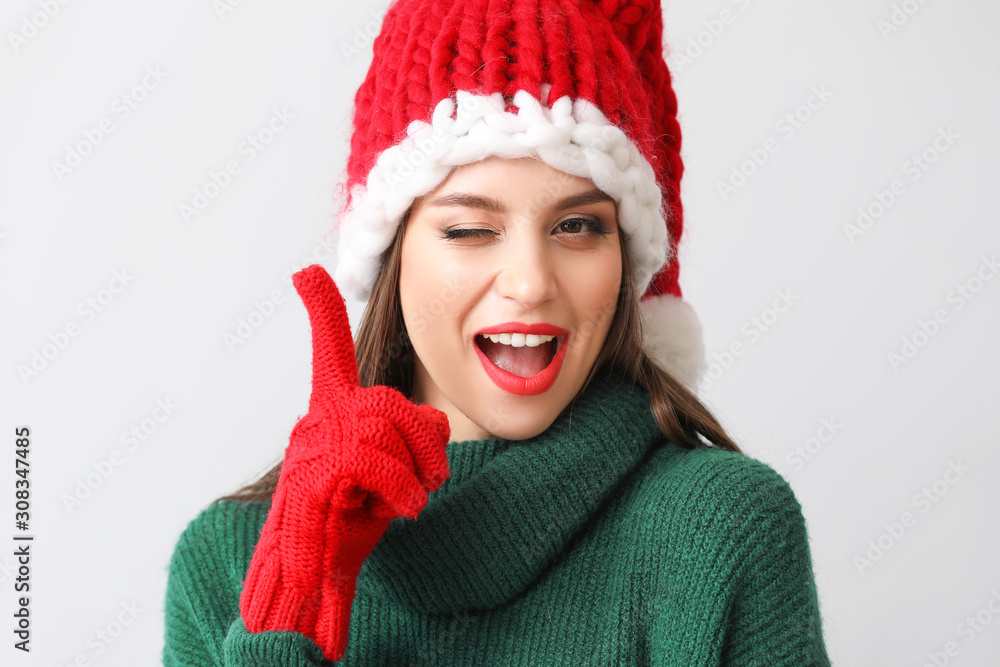 Beautiful young woman in Santa hat and with raised index finger on light background Beautiful young woman in Santa hat and with raised index finger on light background