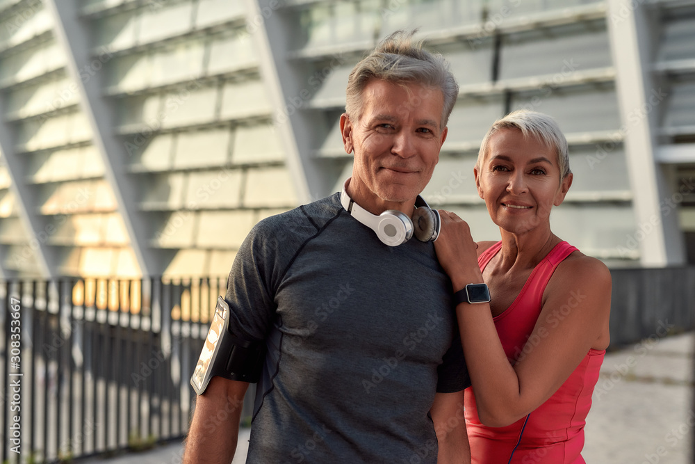 Powerful couple. Portrait of happy middle-aged man and woman in sport clothing looking at camera with smile while standing against stadium outdoors Powerful couple. Portrait of happy middle-aged man and woman in sport clothing looking at camera with smile while standing against stadium outdoors
